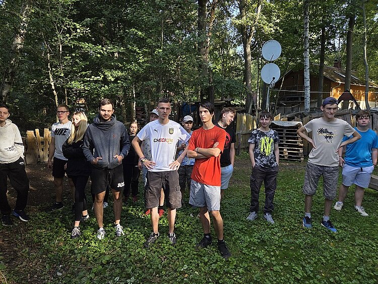 Jugendliche stehen in einem Kreis im Wald, teils mit verschränkten Armen, wirken gespannt.