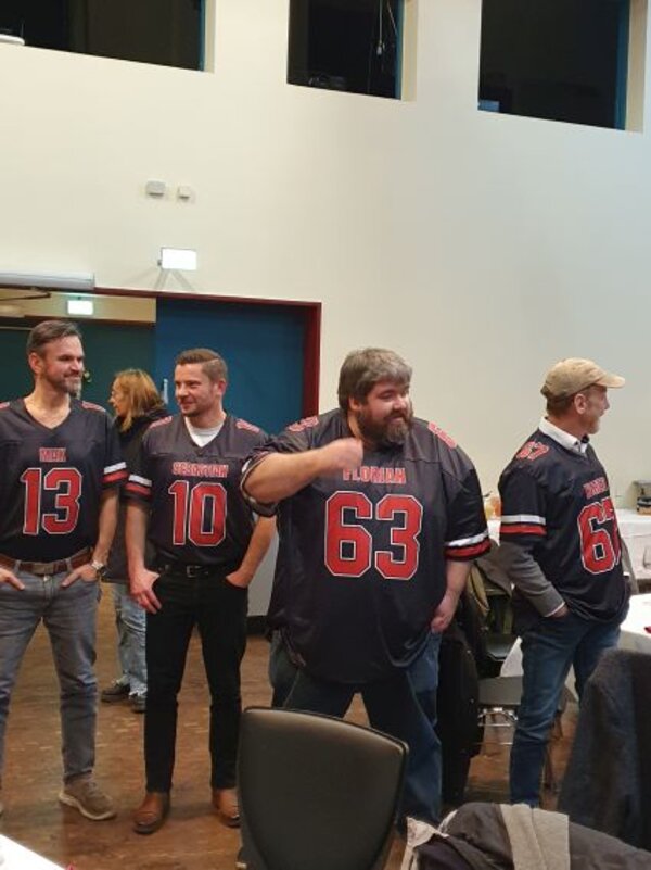 Vier Männer stehen nebeneinander in einem Saal und tragen dunkelblaue Football-Trikots mit roten Streifen und großen Rückennummern. Sie wirken gut gelaunt, einer zeigt gestikulierend nach vorne, im Hintergrund sind Tische und weitere Gäste zu sehen.