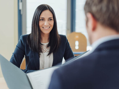 Eine Frau mit langen dunklen Haaren und einem Business-Anzug sitzt lächelnd an einem Tisch und hält einen Ordner. Sie scheint sich in einem Gespräch oder einer Besprechung mit einer anderen Person zu befinden, deren Rücken zu sehen ist. Der Raum ist hell und hat große Fenster, die Tageslicht hereinlassen.