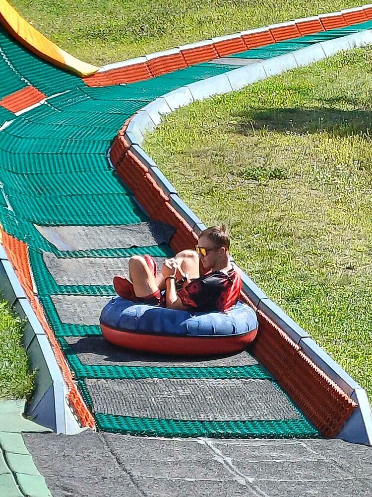 Ein junger Mann rutscht auf einem Ring das letzte Stück einer mit Teppich ausgelegte Rodelbahn herunter. Er trägt eine reflektierende Sonnenbrille.