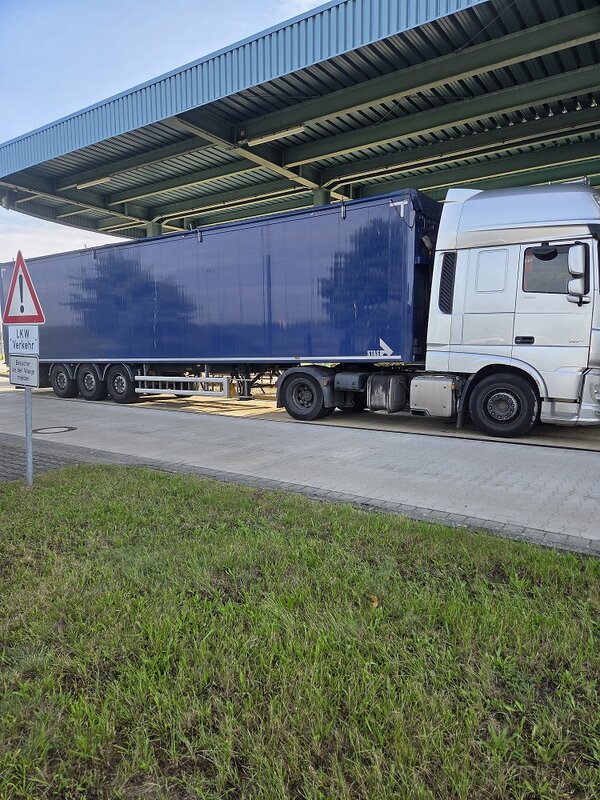 Lastkraftwagen mit blauem Sattelauflieger steht unter einem großen Überdach an einer Laderampe. Links davor befindet sich ein Warnschild „LKW Verkehr“. Im Vordergrund ist eine Rasenfläche zu sehen.