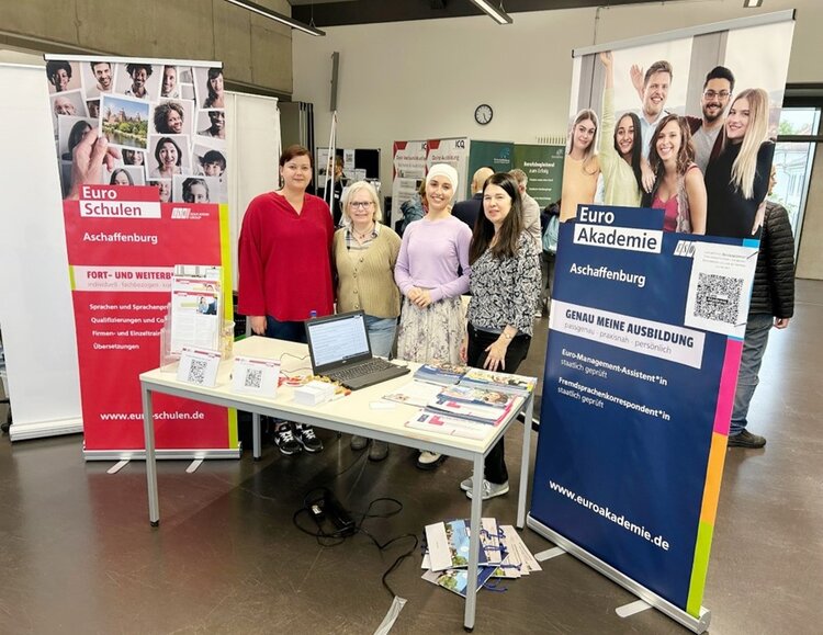 Hinter einem Messestand stehen vier Frauen unterschiedlichen Alters und schauen in die Kamera. Auf dem Tisch steht neben verschiedenen Infobroschüren und Flyern sowie zwei Aufstellern mit QR Codes ein Laptop. Neben dem Tisch stehen Roll-ups der Euro-Schulen (links) und der Euro Akademie (rechts).