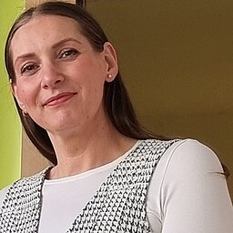 Porträtbild der Dozentin Laura Nicolici, die freundlich in die Kamera lächelt, lange, offene braune Haare zum Mittelscheitel und kleine Ohrstecker sowie ein weißes Longsleeve und eine karrierte Weste darüber trägt.