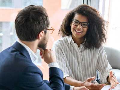 Ein Mann und eine Frau sitzen sich gegenüber und unterhalten sich. Die Frau trägt eine Brille und ein gestreiftes Hemd, während der Mann einen Anzug trägt. Beide wirken entspannt und freundlich, was auf ein angenehmes Gespräch hinweist.