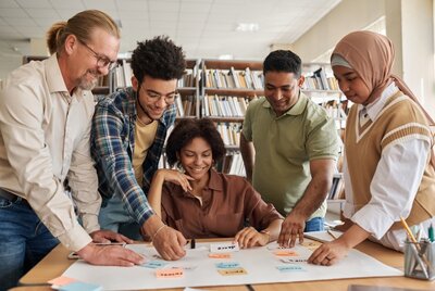 Fünf Personen stehen und sitzen um einen Tisch, auf dem ein großes Blatt Papier mit bunten Haftnotizen liegt. Sie schauen gemeinsam auf die Notizen und zeigen darauf. Im Hintergrund sind Bücherregale mit zahlreichen Büchern zu sehen. Die Personen wirken konzentriert und arbeiten zusammen.