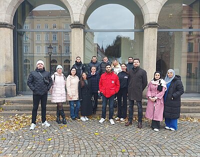 Die weiblichen und männlichen Teilnehmenden des Integrationskurses posieren vor dem Museum Kaisertrutz in Görlitz für ein Gruppenfoto. Alle tragen herbstlich-warme Kleidung, auf dem Kopfsteinpflaster liegt Herbstlaub.