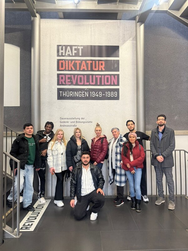 Die Gruppe der Teilnehmenden des Integrationskurses steht und hockt vor einer großen Wand mit einer Ausstellungsgrafik. Die Wand zeigt einen vertikalen Schriftzug mit den Worten 'HAFT', 'DIKTATUR' und 'REVOLUTION' in unterschiedlichen Farben und Schriftgrößen, darunter steht 'THÜRINGEN 1949-1989'. Die Personen sind unterschiedlich gekleidet, einige tragen Jacken oder Mäntel. Im unteren Bereich der Wand befindet sich ein weiterer Textblock. Die Umgebung wirkt modern und ist von Metallgeländern und Treppenstufen eingerahmt.