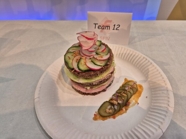 Ein Sandwichturm aus Pumpernickeln, Wurst und Salat mit einer Krönung aus Gurken- und Radieschenscheiben, daneben Gewürzgurken mit Soße auf einem Pappteller; dahinter ein Schildchen mit der Aufschrift "Team 12".