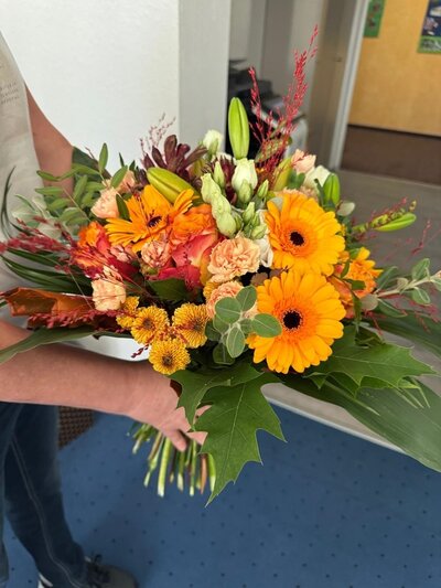 Ein bunter Blumenstrauß mit orangefarbenen Gerbera, gelben und lachsfarbenen Chrysanthemen, weißen und roten Blüten sowie verschiedenen grünen Blättern wird von einer Person im Arm gehalten. Die Person trägt ein kurzärmeliges, helles Oberteil. Im Hintergrund sind ein weißer Wandabschnitt, ein blauer Teppichboden und eine geöffnete Tür zu sehen.