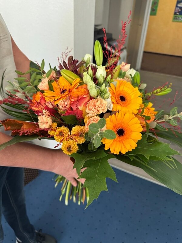 Ein bunter Blumenstrauß mit orangefarbenen Gerbera, gelben und lachsfarbenen Chrysanthemen, weißen und roten Blüten sowie verschiedenen grünen Blättern wird von einer Person im Arm gehalten. Die Person trägt ein kurzärmeliges, helles Oberteil. Im Hintergrund sind ein weißer Wandabschnitt, ein blauer Teppichboden und eine geöffnete Tür zu sehen.