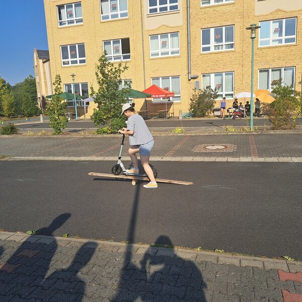 Auf dem Parkplatz vor dem Schulgebäude fährt eine Schülerin mit einem Roller über eine Wippe, die aus einem Stück Holz und einem langen Brett improvisiert ist. Vorne im Bild kann man die Schatten der Zuschauer auf dem Asphalt erkennen. Im Hintergrund sind entlang des Schulgebäudes Sonnenschirme und Bänke aufgestellt.