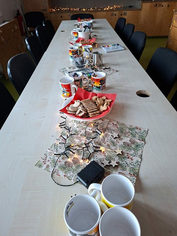 Langer Tisch im Aufenthaltsraum, festlich gedeckt mit Bechern, Keksen, Servietten und Lichterkette für eine Adventsrunde.
