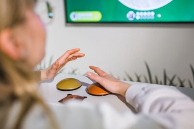 Eine Person sitzt vor einem Bildschirm und hält ihre Hände über einen gelben und einen orangefarbenen Buzzer. Sie scheint an einem digitalen Quiz teilzunehmen.
