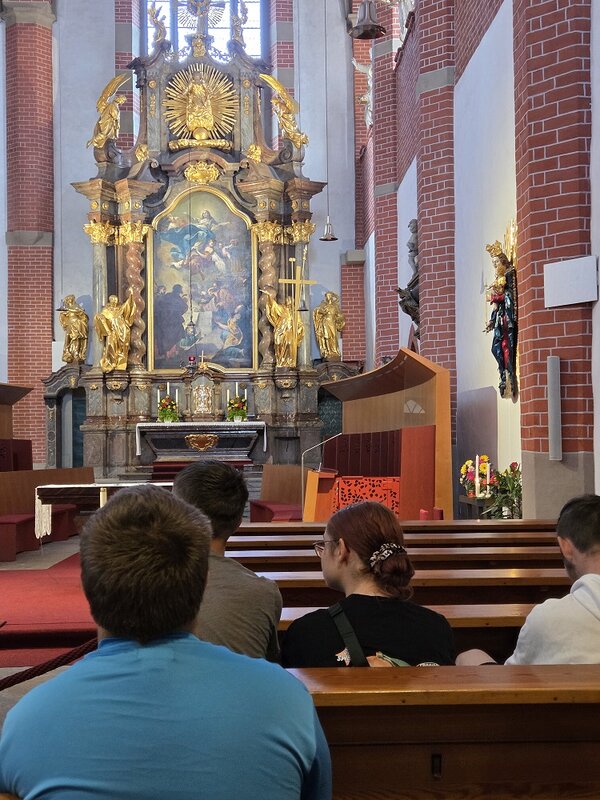 Die Rückansicht von Personen, die in einer Kirche in den Bänken sitzen. Im Hintergrund ist ein prächtiger, goldverzierter Altar mit einem Gemälde und Skulpturen zu sehen. Die Kirche hat rote Ziegelwände und helle Fenster.