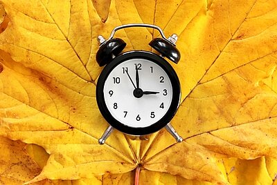 Die Uhr umstellen Ein schwarzer Wecker, der auf drei Uhr steht, liegt auf herbstlich gelbem Ahornblatt.