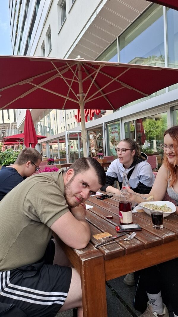 Vier Mitglieder der Pflegefachklasse sitzen entspannt an einem Holztisch im Außenbereich eines Restaurants. Im Vordergrund lehnt sich ein junger Mann mit freundlichem Blick zur Kamera. Um ihn herum unterhalten sich die anderen, während sie ihr Essen genießen. Große rote Sonnenschirme spenden Schatten, im Hintergrund sind moderne Gebäude mit Glasfassaden zu sehen – ein Moment der Erholung mitten in der Stadt.