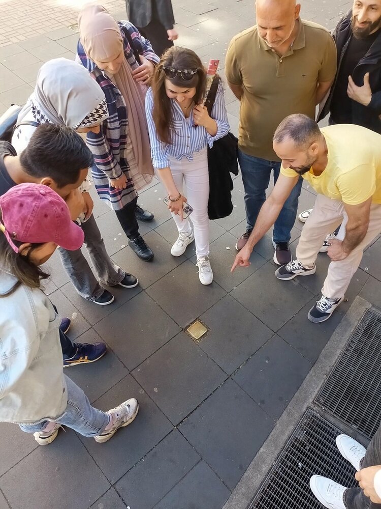 Die Teilnehmerinnen und Teilnehmer des Orientierungskurses an den Euro-Schulen Mainz sind über einen Stolperstein auf dem Gehweg gebeugt und lesen die Inschrift.