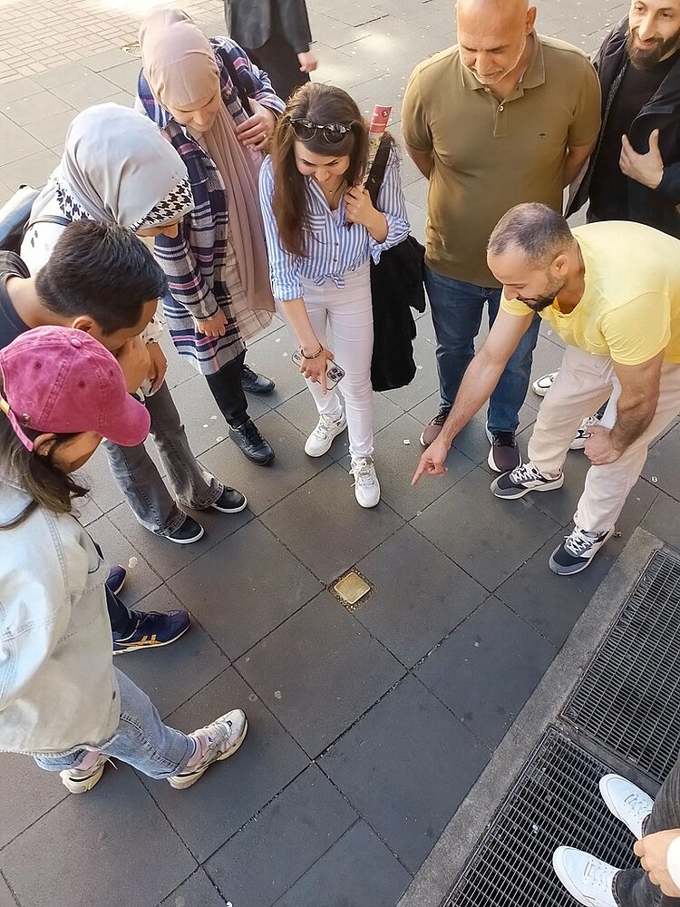 Der Orientierungskurs – Ein wichtiger Baustein in den Integrationskursen Die Teilnehmerinnen und Teilnehmer des Orientierungskurses an den Euro-Schulen Mainz sind über einen Stolperstein auf dem Gehweg gebeugt und lesen die Inschrift.