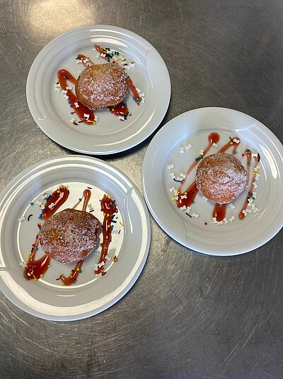 Kein Karneval in Bielefeld Drei weiße Teller mit jeweils einem mit Zucker bestreuten Krapfen (Berliner) sind auf einem Edelstahltisch angerichtet. Die Teller sind mit roten Soßenstreifen sowie bunten Zuckerstreuseln dekoriert.