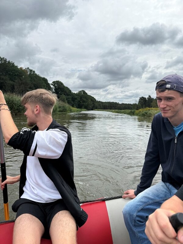 Zwei junge Männer in einem roten Schlauchboot auf einem Fluss. Einer von ihnen paddelt, während der andere nach vorne schaut. Der Himmel ist bewölkt, und im Hintergrund sind Bäume und Sträucher zu erkennen.