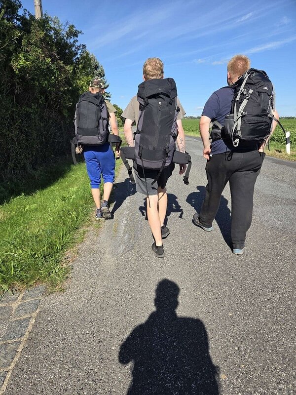 Drei Wanderer mit großen Rucksäcken gehen auf einer asphaltierten Straße durch eine ländliche Gegend. Die Sonne scheint hell, und die Schatten der Personen sind deutlich auf dem Boden zu sehen. Im Hintergrund sind Felder und Bäume zu erkennen.
