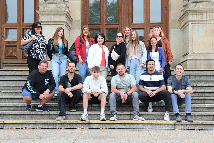 Unser Integrationskurs besucht Leipzig Gruppenfoto von 15 Personen auf einer Treppe vor einem historischen Gebäude mit Holztüren.