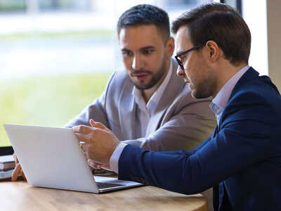Zwei Männer in einem professionellen Umfeld sitzen an einem Tisch und arbeiten gemeinsam an einem offenen Laptop. Der Mann im Vordergrund trägt ein dunkles Sakko und eine Brille, während der andere Mann im Hintergrund ein hellgraues Sakko trägt. Beide wirken konzentriert und scheinen sich über den Inhalt auf dem Bildschirm auszutauschen. Der Hintergrund zeigt ein helles Büro oder Besprechungszimmer mit großen Fenstern.