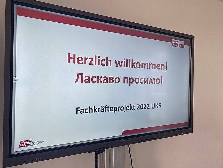 Freude am Erfolg beim Integrationskurs Ein großer Monitor zeigt in roten Buchstaben Herzlich Willkommen an, darunter steht selbiges in ukrainisch.