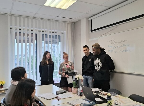 Zwei weibliche und zwei männliche Teilnehmende stehen vorn am Whiteboard und tragen ihren Präsentation vor.