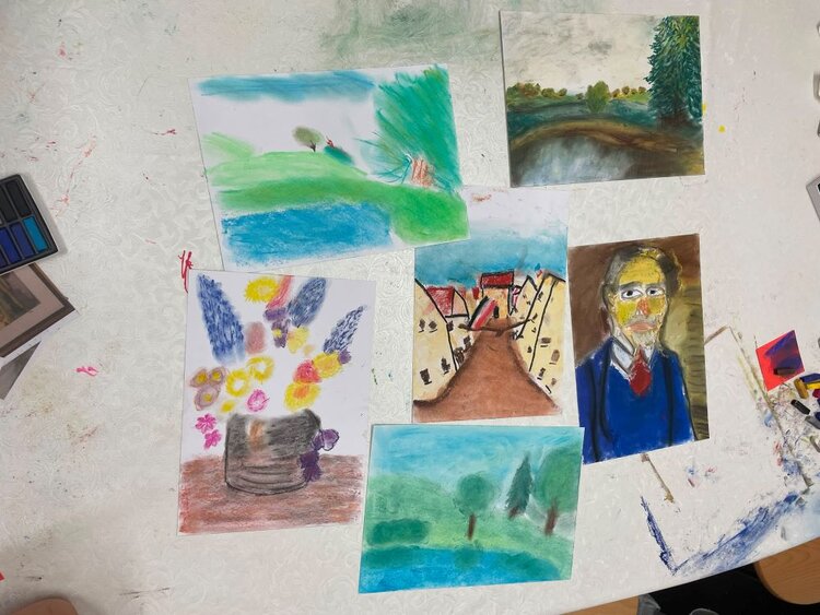 Mehrere bunte Zeichnungen und Gemälde liegen auf einem Tisch. Die Bilder zeigen verschiedene Motive wie Landschaften, Porträts und abstrakte Kompositionen. Im Hintergrund sind Malutensilien wie Pinsel, Farben und Papier zu sehen. 