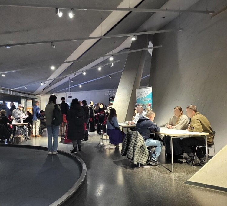 Bewerbungsgespräche beim Job-Speed-Dating der Euro-Schulen Märkischer Kreis in der Phänomenta in Lüdenscheid: Mehrere Arbeitgeber sitzen an Tischen entlang einer modernen Betonwand im Ausstellungsraum. Gegenüber stehen oder sitzen Besucherinnen und Besucher, die auf Gespräche warten. Die Veranstaltung ist gut besucht, und im Raum herrscht eine konzentrierte, aber offene Atmosphäre. Informationsbanner verschiedener Unternehmen stehen im Hintergrund, während sich Teilnehmende aktiv miteinander austauschen.