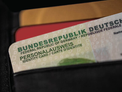Ein Ausschnitt eines deutschen Personalausweises, der die Aufschrift 'Bundesrepublik Deutschland' in deutscher, englischer und französischer Sprache zeigt. Der Ausweis liegt auf einem schwarzen Untergrund, teilweise überlappt von einer roten und einer goldfarbenen Karte.