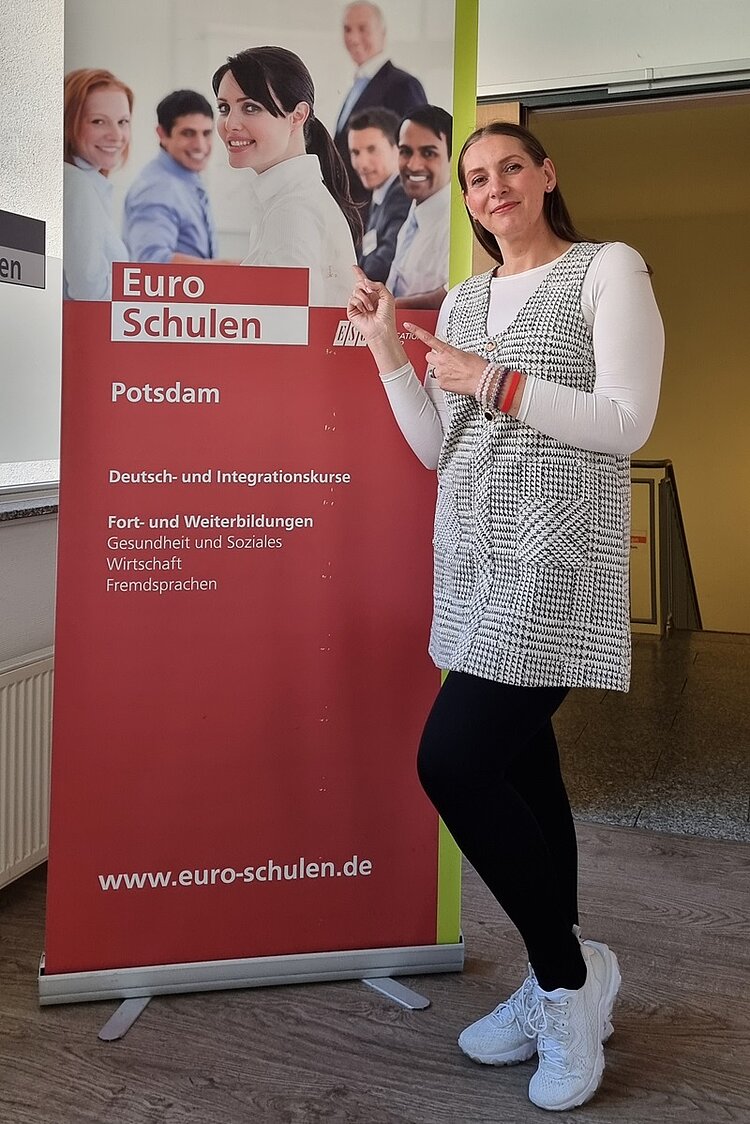 Frau Laura Nicolici posiert lächelnd als neue Kollegin vor einem großen Roll-up der Euro-Schulen Potsdam, auf das sie mit beiden Zeigefindern zeigt. Sie trägt weiße Sneaker, eine dunkle Leggings, ein weißes Longsleeve und darüber eine schwarz weiß karrierte Tunika.