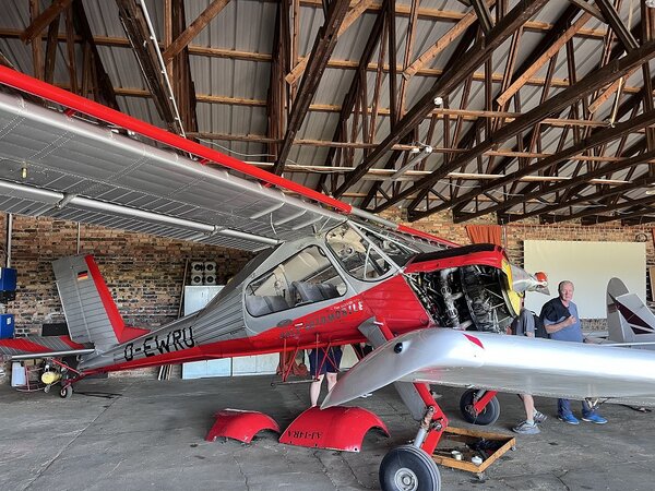 Rot-silbernes Motorflugzeug in einem Hangar mit geöffnetem Motorbereich; daneben stehen mehrere Personen.