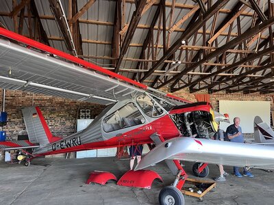 Rot-silbernes Motorflugzeug in einem Hangar mit geöffnetem Motorbereich; daneben stehen mehrere Personen.