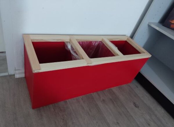 Ein rechteckiger Mülltrennungsbehälter aus Holz mit roten Seitenwänden, bestehend aus drei Fächern. In der Mitte ist ein Fach mit einem Müllbeutel ausgestattet, während die anderen beiden Fächer leer sind. Der Behälter steht auf einem Parkettboden und ist vor einer weißen Wand platziert.