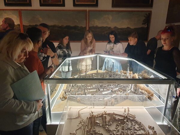 Eine Gruppe von Menschen steht um ein detailliertes Modell der Stadt Zittau, das in einer Glasvitrine ausgestellt ist. Die Personen betrachten das Modell aufmerksam, während sie sich in einem Raum mit historischen Gemälden an den Wänden befinden.