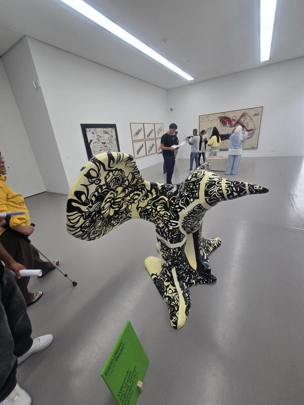 In einem großen Ausstellungsraum steht eine auffällig schwarz-creme gemusterte Skulptur im Vordergrund; im Hintergrund betrachten Teilnehmende und Besucher*innen Zeichnungen und großformatige Kunstwerke an den Wänden.