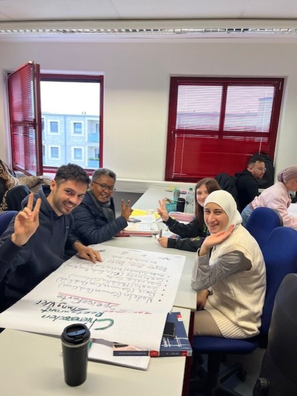 Vier Teilnehmende des Berufssprachkurses sitzen an ihrer Projektarbeit und lächeln in die Kamera.