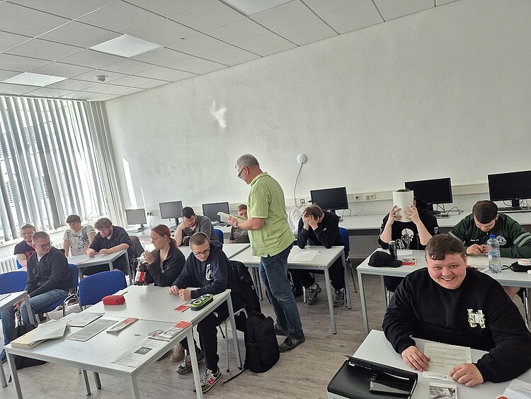 Eine Gruppe von Schülern sitzt in einem Computerraum an weißen Tischen. Einige unterhalten sich oder arbeiten mit Arbeitsblättern. Ein Lehrer in grünem Hemd steht in der Mitte des Raumes und spricht mit einer Schülergruppe. Die Stimmung wirkt konzentriert und locker.