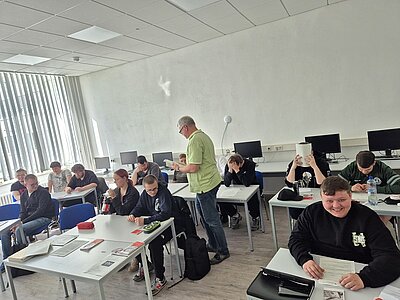 Eine Gruppe von Schülern sitzt in einem Computerraum an weißen Tischen. Einige unterhalten sich oder arbeiten mit Arbeitsblättern. Ein Lehrer in grünem Hemd steht in der Mitte des Raumes und spricht mit einer Schülergruppe. Die Stimmung wirkt konzentriert und locker.