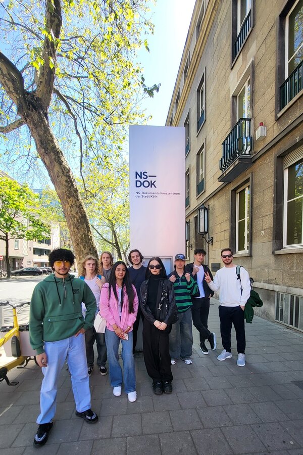 Die Gruppe des Jugendintegrationskurses steht auf einem Gehweg vor einem hohen, rechteckigen Schild mit der Aufschrift 'NS-DOK' und weiteren kleineren Textzeilen. Im Hintergrund ist ein mehrstöckiges Gebäude mit großen Fenstern und Balkonen zu sehen. Ein großer Baum mit ausladenden Ästen spendet Schatten. Die Personen tragen Freizeitkleidung und blicken in Richtung Kamera. Das Wetter ist sonnig, der Himmel blau.