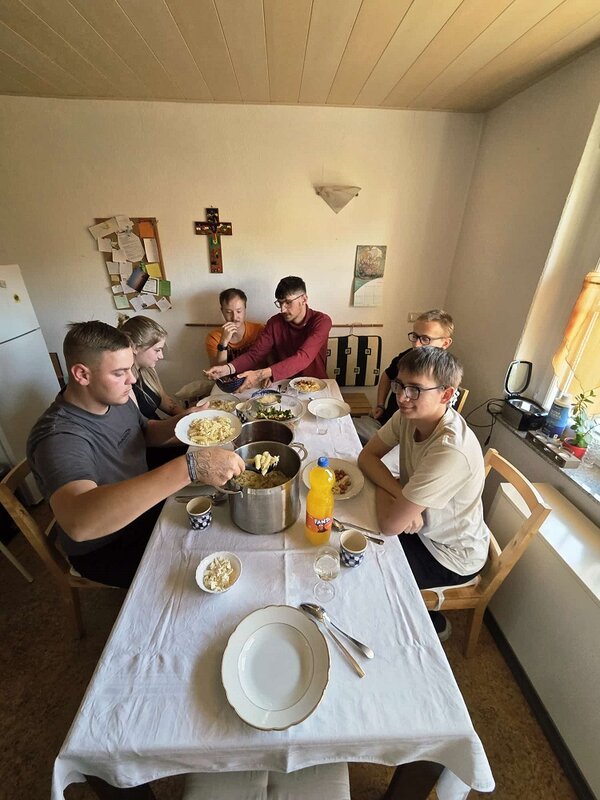 Eine Gruppe junger Menschen sitzt gemeinsam an einem gedeckten Tisch und isst eine warme Mahlzeit in einer einfachen Unterkunft.