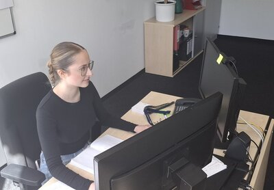 Auszubildende der Euro-Schulen und der Euro Akademie Berlin, Chiara Postaru, sitzt an einem Schreibtisch vor dem Computerbildschirm, sie trägt ein dunkles Oberteil, eine Brille und die Haare zusammengebunden zu einem Zopf. 