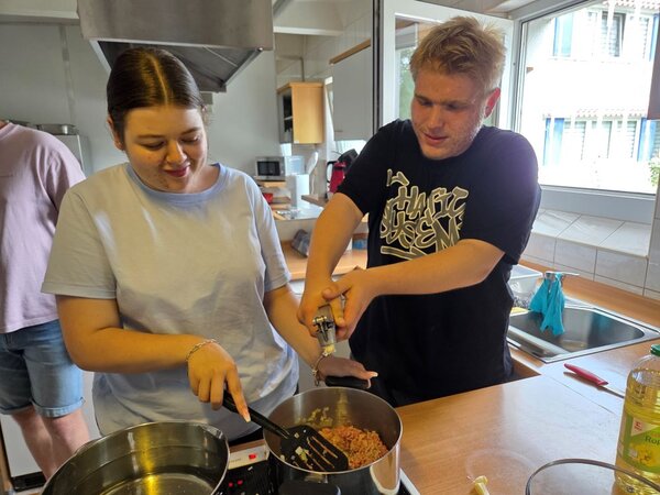 Zwei Auszubildende stehen nebeneinander an einem Herd in einer Schulküche. Eine junge Frau rührt mit einem Pfannenwender in einem Topf mit Hackfleisch, während ein junger Mann mit beiden Händen eine Knoblauchpresse bedient. Beide wirken konzentriert und gut gelaunt.