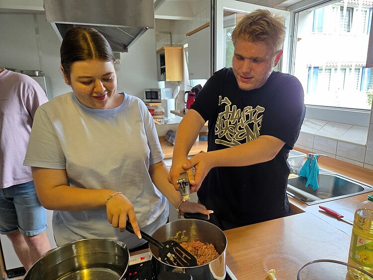 Zwei Auszubildende stehen nebeneinander an einem Herd in einer Schulküche. Eine junge Frau rührt mit einem Pfannenwender in einem Topf mit Hackfleisch, während ein junger Mann mit beiden Händen eine Knoblauchpresse bedient. Beide wirken konzentriert und gut gelaunt.