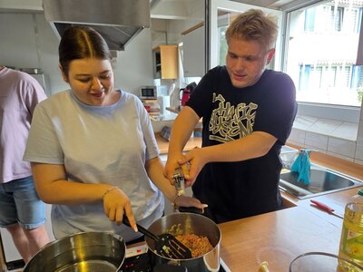 Zwei Auszubildende stehen nebeneinander an einem Herd in einer Schulküche. Eine junge Frau rührt mit einem Pfannenwender in einem Topf mit Hackfleisch, während ein junger Mann mit beiden Händen eine Knoblauchpresse bedient. Beide wirken konzentriert und gut gelaunt.