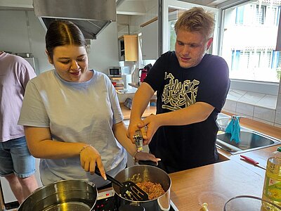 Zwei Auszubildende stehen nebeneinander an einem Herd in einer Schulküche. Eine junge Frau rührt mit einem Pfannenwender in einem Topf mit Hackfleisch, während ein junger Mann mit beiden Händen eine Knoblauchpresse bedient. Beide wirken konzentriert und gut gelaunt.