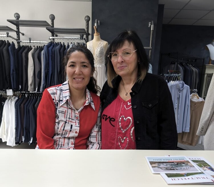 Zwei Frauen sitzen an einem weißen Tisch und lächeln in die Kamera. Im Hintergrund sind Kleiderstangen mit Anzügen und eine Schneiderpuppe mit einem Brautkleid zu sehen. Vorne rechts auf dem Tisch liegen ein paar Zeitschriften.