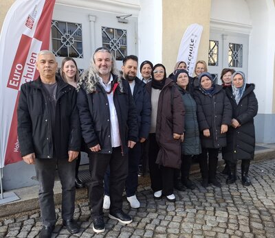 Eine Gruppe von Kursteilnehmenden steht auf einem gepflasterten Platz vor einem Gebäude mit weißen Wänden und verzierten Fenstern. Alle tragen dunkle Winterjacken und stehen nebeneinander. Im Hintergrund sind zwei weiße Fahnen mit roten und schwarzen Schriftzügen zu sehen.